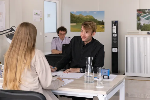 Zwei Menschen sitzen an einem Schreibtisch in einem Büro, sie scheinen ein Gespräch zu führen. Im Hintergrund arbeitet eine Person am Laptop.